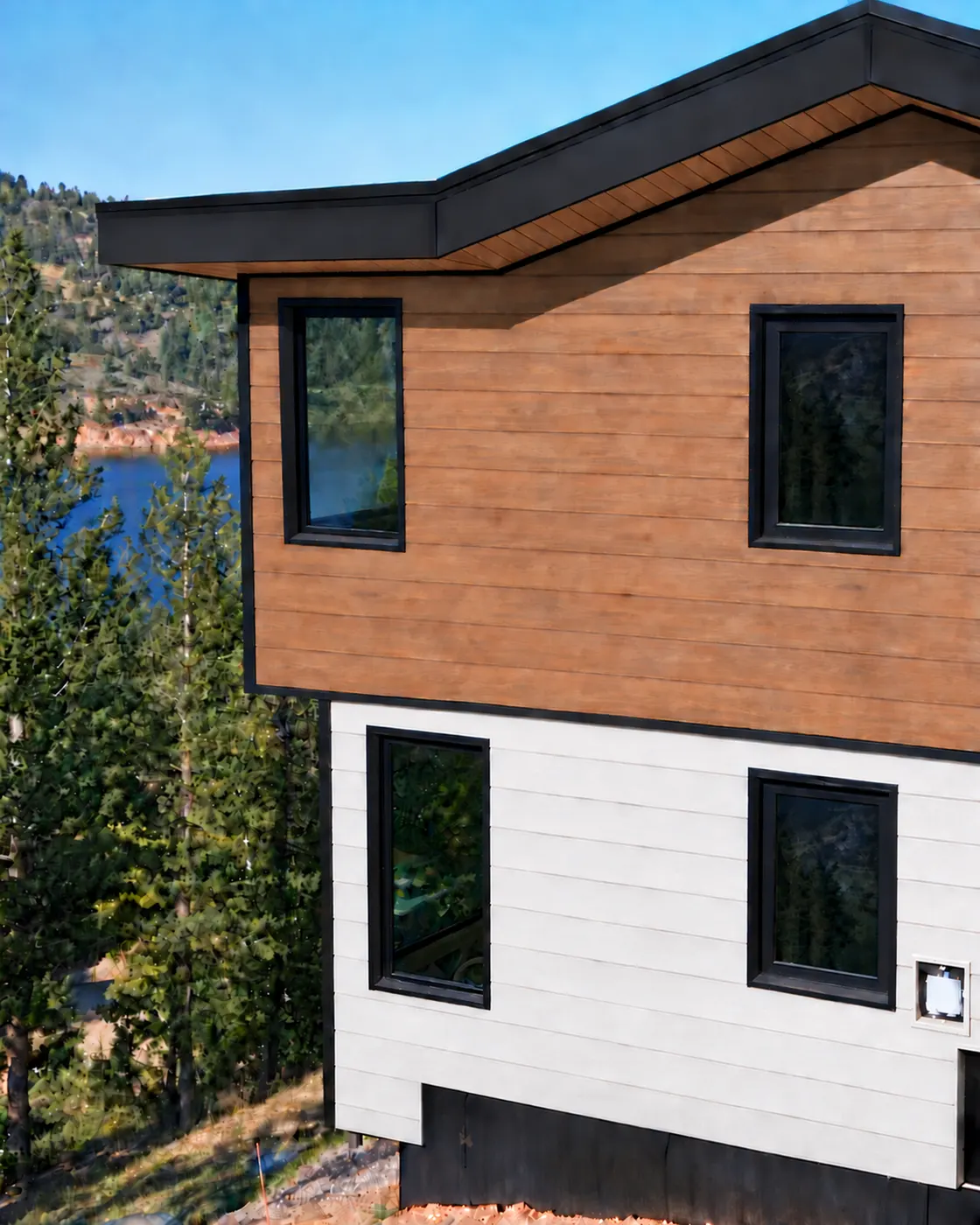 Upper-storey wall in wood-look siding above light horizontal siding, with black-framed windows and pines along a lake below. EDCO hickory and glacier white metal siding, with Panda windows and doors.