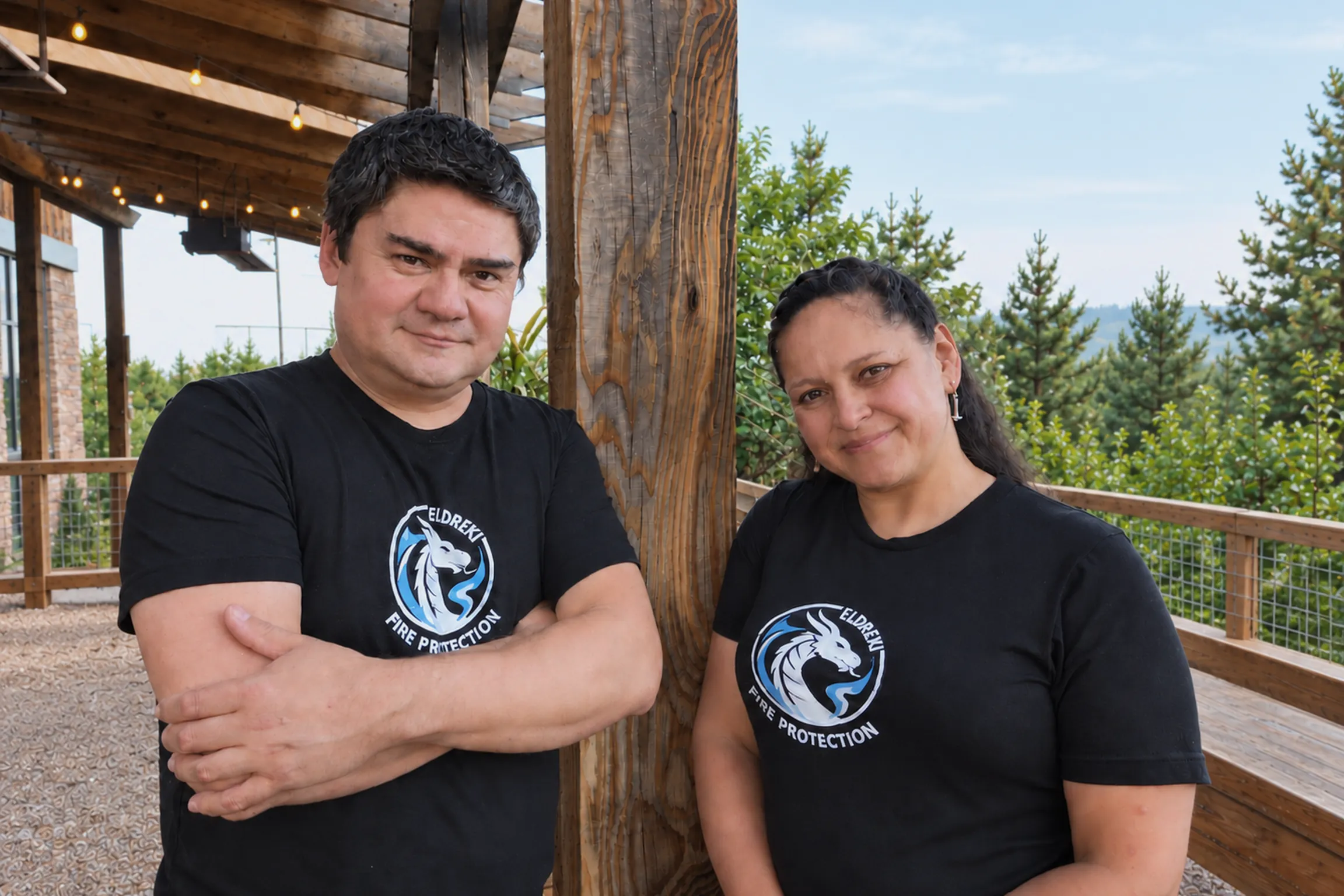 Two people in black Eldreki Fire Protection shirts, standing on a covered deck with a heavy timber post between them and conifers on the hillside behind.